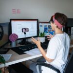 A person sits in an office chair at a desk with multiple monitors displaying work. In front of the monitors, a desktop tripod holds a front-facing smartphone. They are gesturing while they record a video. They are wearing a white tee shirt and a red and blue scarf over their short hair. The photo accompanies a blog post about workplace TikTok trends, such as quiet quitting, acting your wage, and task masking, and the relationship between personality and employee engagement.