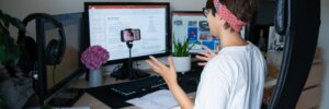 A person sits in an office chair at a desk with multiple monitors displaying work. In front of the monitors, a desktop tripod holds a front-facing smartphone. They are gesturing while they record a video. They are wearing a white tee shirt and a red and blue scarf over their short hair. The photo accompanies a blog post about workplace TikTok trends, such as quiet quitting, acting your wage, and task masking, and the relationship between personality and employee engagement.