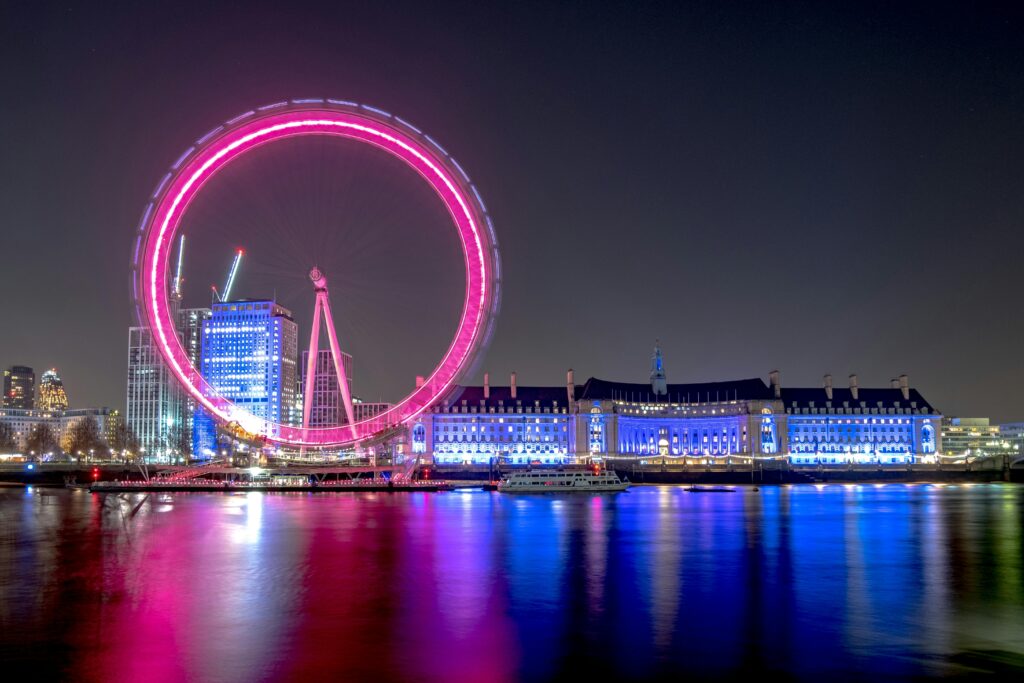In this night view of the London skyline, the London Eye is illuminated in bright pink neon lights, and the historic County Hall building is lit in blue. The vibrant atmosphere is reflected in the River Thames in the foreground.