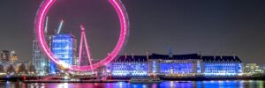 In this night view of the London skyline, the London Eye is illuminated in bright pink neon lights, and the historic County Hall building is lit in blue. The vibrant atmosphere is reflected in the River Thames in the foreground.
