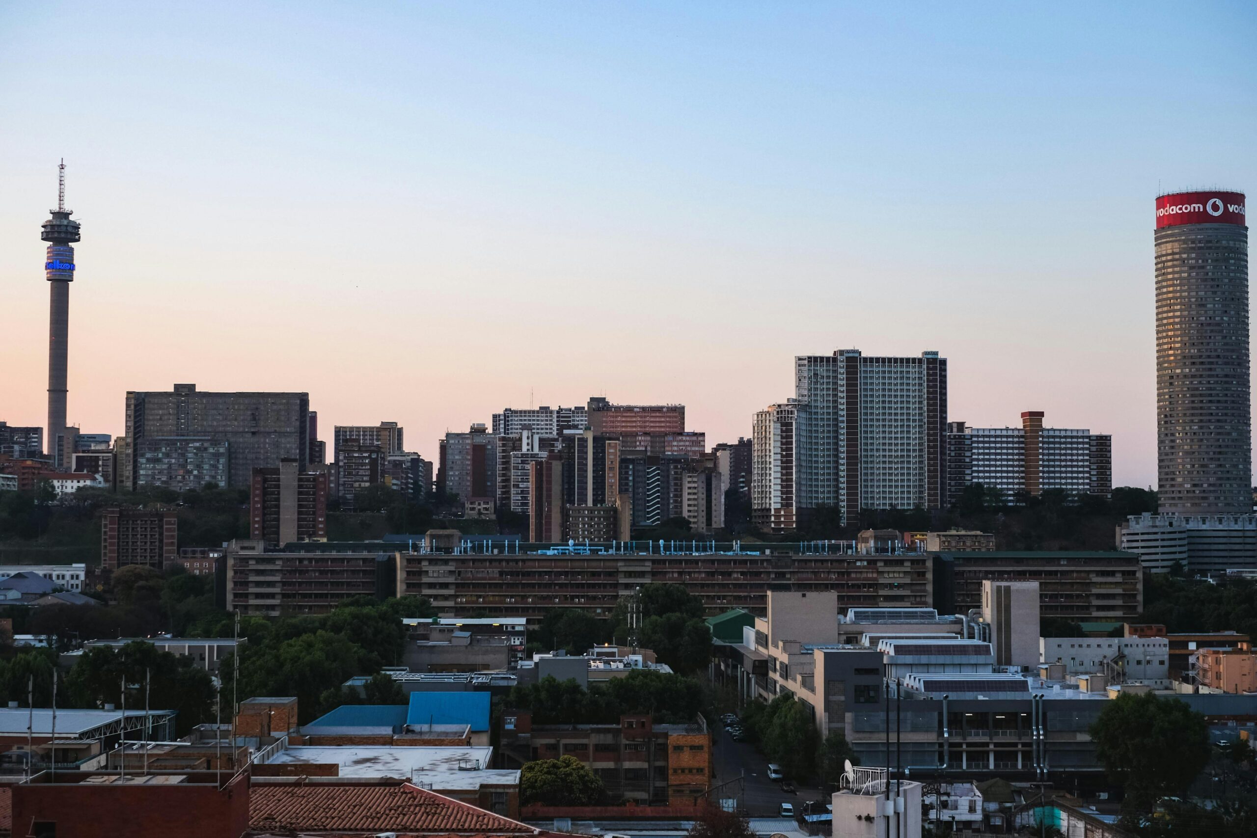 A view of downtown Johannesburg, South Africa, at dusk shows a dense urban cityscape with multiple high-rise buildings and towers under a sky turning sunset pink and orange near the horizon. The image accompanies an article highlighting personality trends of South African leaders.