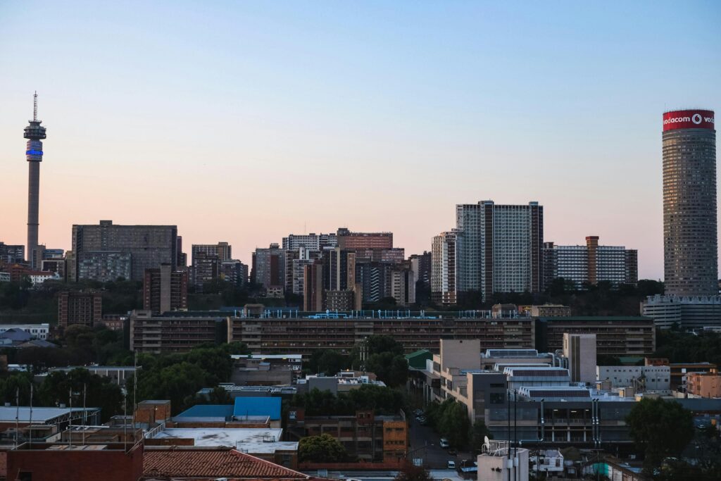 A view of downtown Johannesburg, South Africa, at dusk shows a dense urban cityscape with multiple high-rise buildings and towers under a sky turning sunset pink and orange near the horizon. The image accompanies an article highlighting personality trends of South African leaders.
