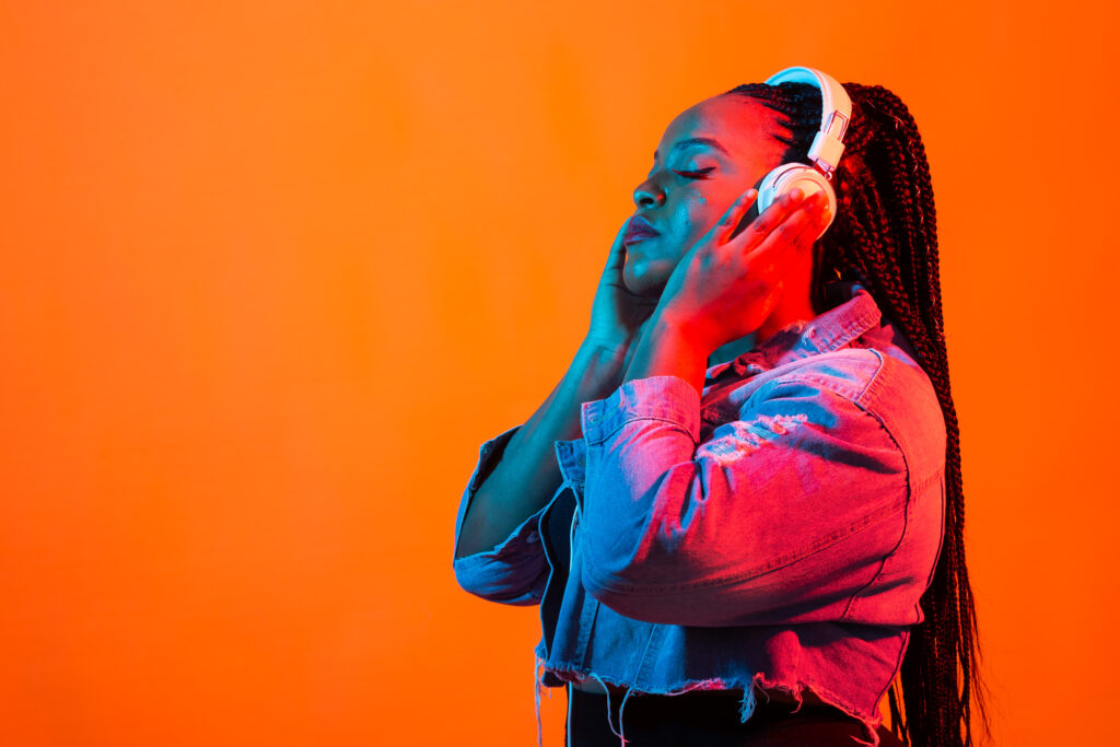 A young Black woman with long braided hair, white headphones, and a denim jacket closes her eyes, appearing deeply absorbed in the music against a vibrant orange background with neon lighting. The image accompanies an article about the connection between music and psychology.