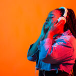 A young Black woman with long braided hair, white headphones, and a denim jacket closes her eyes, appearing deeply absorbed in the music against a vibrant orange background with neon lighting. The image accompanies an article about the connection between music and psychology.