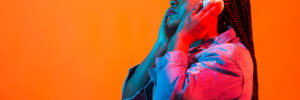 A young Black woman with long braided hair, white headphones, and a denim jacket closes her eyes, appearing deeply absorbed in the music against a vibrant orange background with neon lighting. The image accompanies an article about the connection between music and psychology.