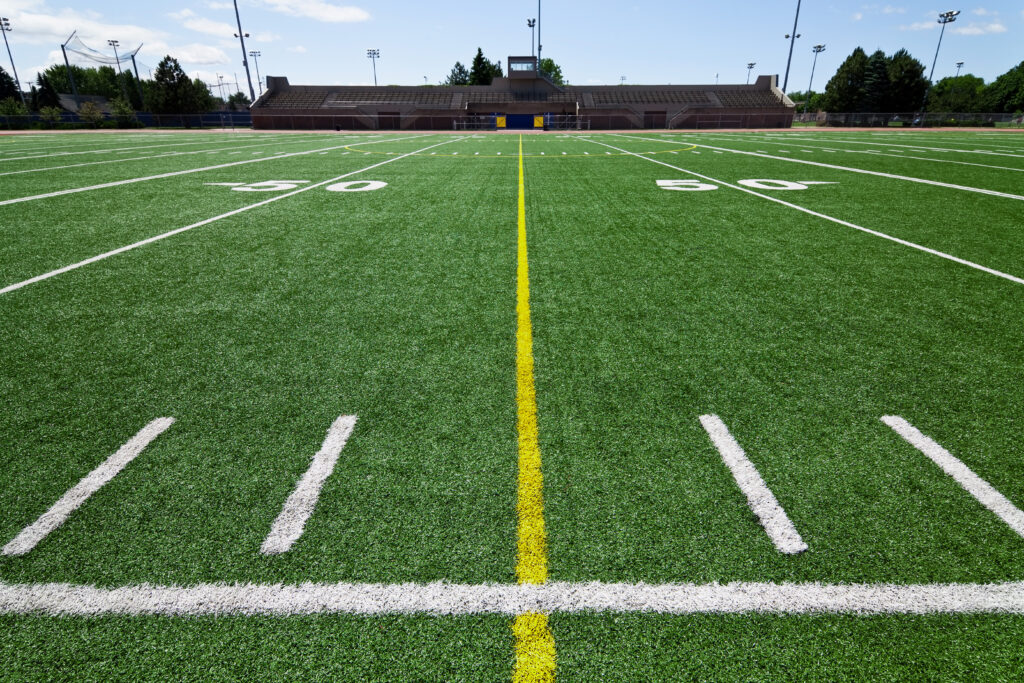 An American football field shows the 50-yard line with white hash marks, white yard numbers, and a yellow center stripe on green artificial turf. The image accompanies an article about predicting athletic success with personality assessment.
