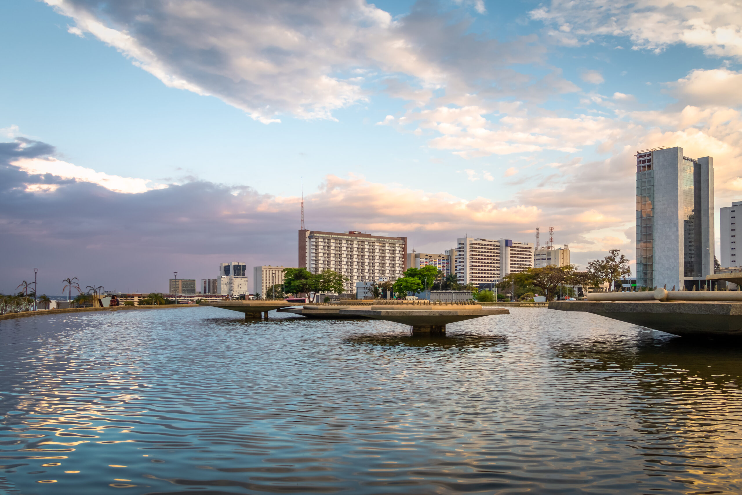 A modern concrete fountain sits in the center of a reflective pool at Burle Marx Garden Park in Brasília, Brazil, with the distinctive skyline of the federal district outlined against a dramatic cloudy sky. The image accompanies an article about emergent personality trends of Brazilian leaders.