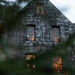 Golden lights illuminate the windows of an old stone building with an eerie, haunted ambiance, viewed from a distance through out-of-focus leaves. The image accompanies an article about why people believe in the paranormal.