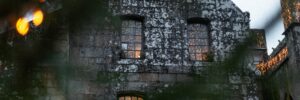 Golden lights illuminate the windows of an old stone building with an eerie, haunted ambiance, viewed from a distance through out-of-focus leaves. The image accompanies an article about why people believe in the paranormal.
