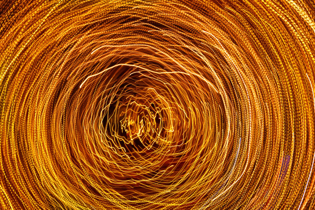 A long-exposure photograph depicts counterclockwise spiraling lines of gold and white light against a black background. The image suggests a creative yet unfocused state of mind, a metaphor for the dark side of creativity, when imagination derails performance, or the Hogan Development Survey Imaginative scale derailer.