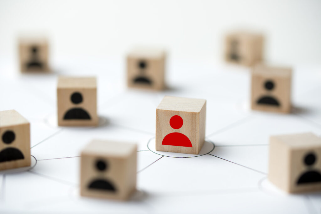 This leadership selection visual metaphor depicts wooden blocks arranged in a network pattern with one red silhouette standing out among black silhouettes. It accompanies an article about new requirements for executive assessment for federal agencies.