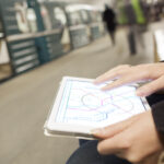A tablet displaying a metropolitan train map is held in a person's lap. They are light-skinned and manipulating the map with their hands. Their face is out of the frame. An out-of-focus train is visible in the background. The photo accompanies a blog post about a leadership coaching framework called the mirror and the map.