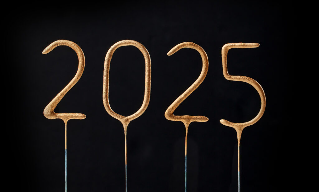 The year 2025 is displayed using four unlit golden sparklers, each digit individually mounted on a thin stick against a black background. The image accompanies an article about the 2025 derailers of the year.
