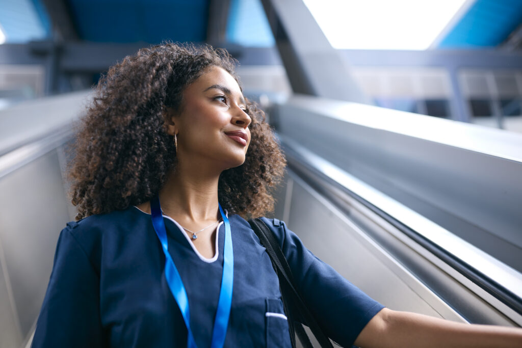 A woman with curly hair wearing blue medical scrubs in a modern office corridor is looking upward with confident expression, representing a technical expert receiving leadership development.