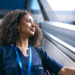 A woman with curly hair wearing blue medical scrubs in a modern office corridor is looking upward with confident expression, representing a technical expert receiving leadership development.