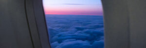 The view through an airplane window shows an expanse of blue-tinted clouds that resemble ocean waves below a pink-and-purple sunset sky along the horizon.