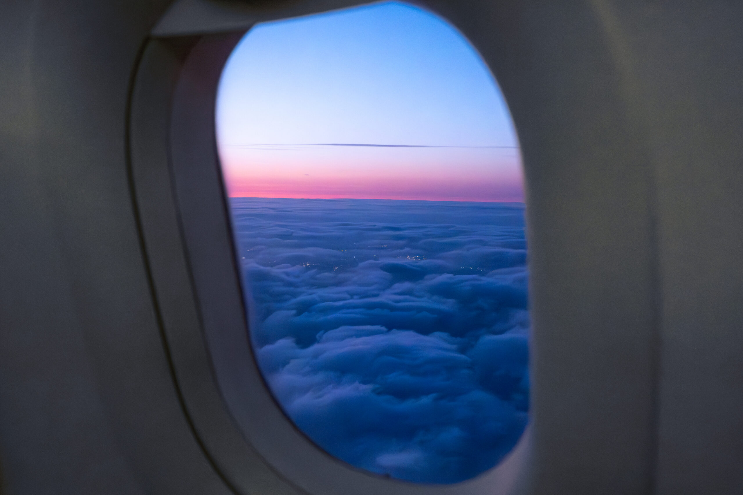 The view through an airplane window shows an expanse of blue-tinted clouds that resemble ocean waves below a pink-and-purple sunset sky along the horizon.