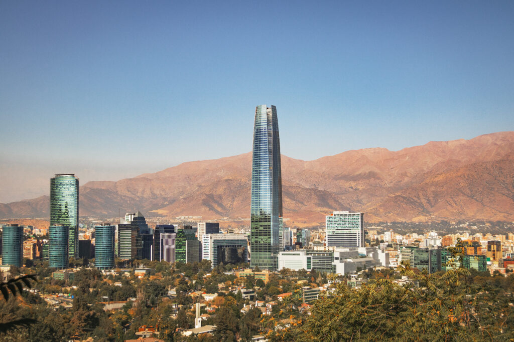 An aerial view of Santiago’s modern skyline features the Costanera Center skyscraper and glass office buildings against the backdrop of the arid Andes Mountains under a clear blue sky. The image accompanies an article about leadership trends of Chilean executives.
