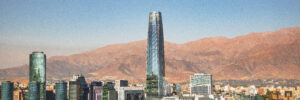 An aerial view of Santiago’s modern skyline features the Costanera Center skyscraper and glass office buildings against the backdrop of the arid Andes Mountains under a clear blue sky. The image accompanies an article about leadership trends of Chilean executives.