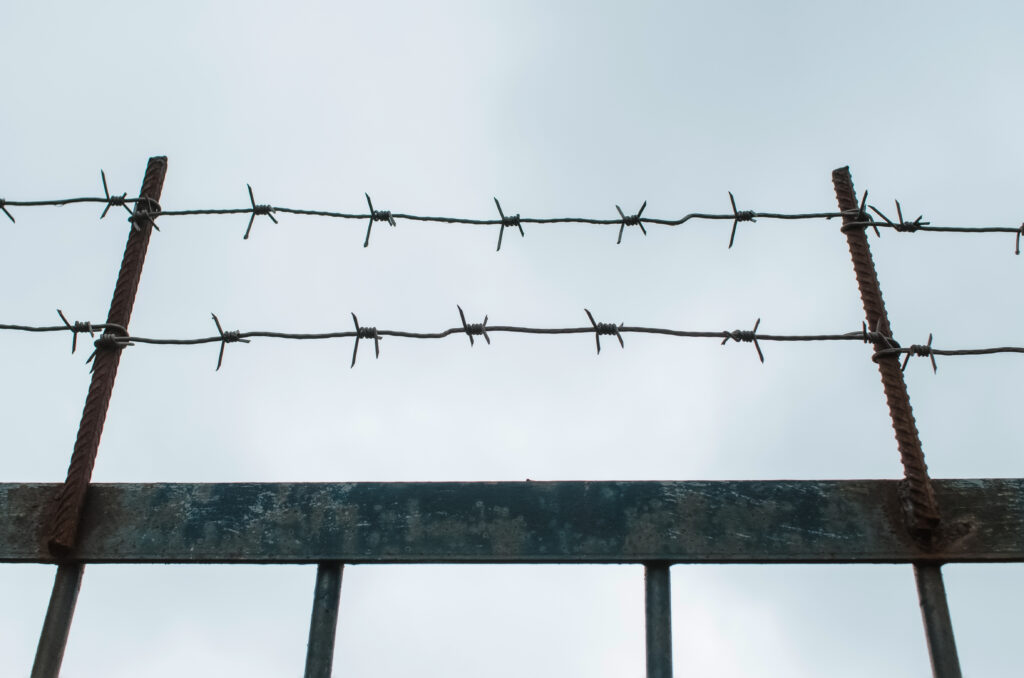 A close-up photo of a barbed-wire fence against a dusky sky, conceptualizing the idea of confinement, imprisonment, etc., to accompany a blog about the dark side of personality and evil personality among authoritarian leaders