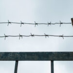 A close-up photo of a barbed-wire fence against a dusky sky, conceptualizing the idea of confinement, imprisonment, etc., to accompany a blog about the dark side of personality and evil personality among authoritarian leaders