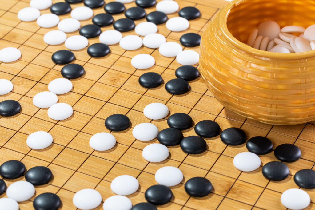 Black and white stones are placed across a wooden Go board, with a woven bamboo bowl of white stones resting on the right side of the board. The image suggests an AI system that plays the board game Go and accompanies an article about personality psychology in an AI-driven workplace.
