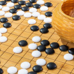 Black and white stones are placed across a wooden Go board, with a woven bamboo bowl of white stones resting on the right side of the board. The image suggests an AI system that plays the board game Go and accompanies an article about personality psychology in an AI-driven workplace.