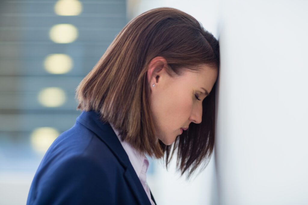 A woman with a light skin tone and straight, shoulder-length brown hair, dressed in professional attire, leans her forehead against a wall with her eyes downcast. The image accompanies an article about the global manager engagement crisis.