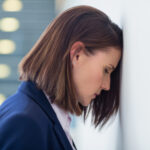 A woman with a light skin tone and straight, shoulder-length brown hair, dressed in professional attire, leans her forehead against a wall with her eyes downcast. The image accompanies an article about the global manager engagement crisis.