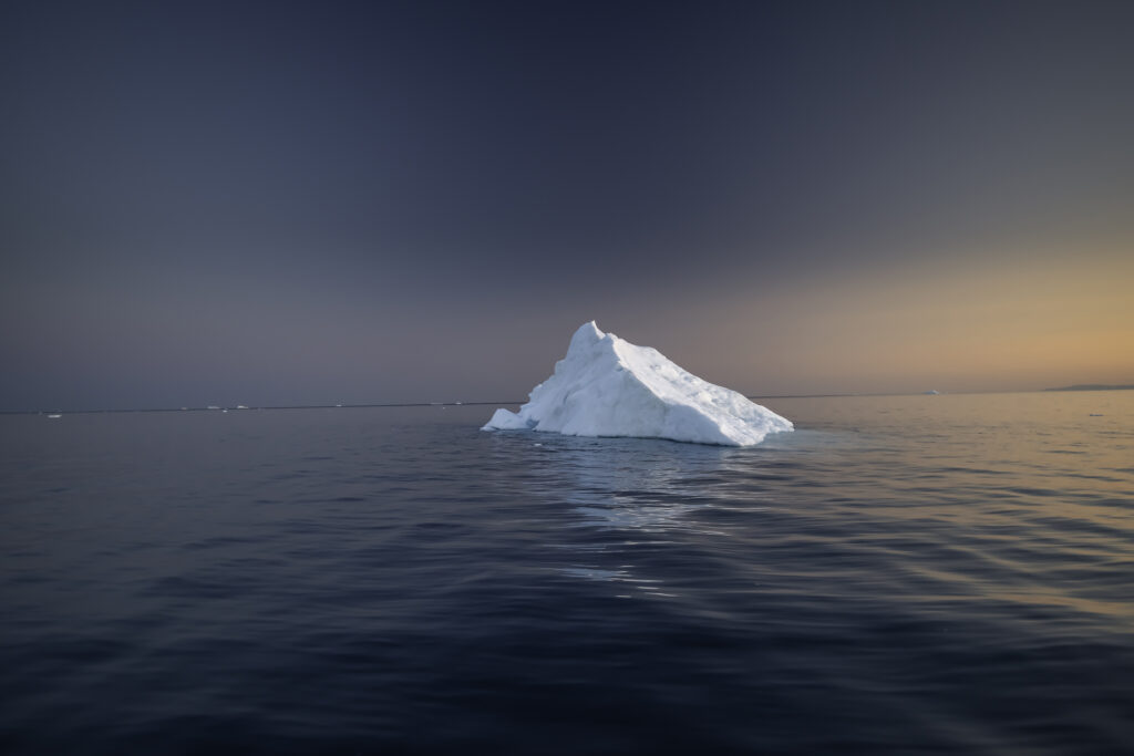 A white pyramid-shaped iceberg floats in dark open water at dusk with low golden light on the right side of the horizon and an overcast sky above. The image accompanies an article about the secret life of organizations.
