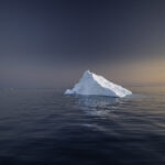 A white pyramid-shaped iceberg floats in dark open water at dusk with low golden light on the right side of the horizon and an overcast sky above. The image accompanies an article about the secret life of organizations.