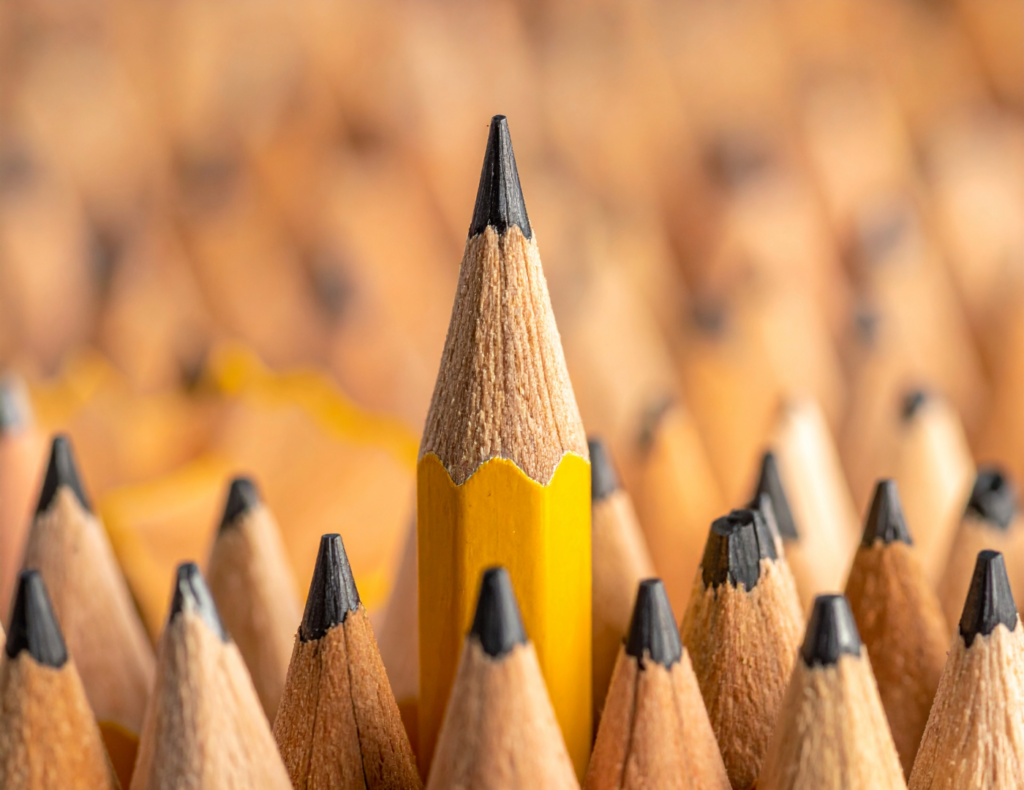 One sharpened yellow pencil rises above a field of near-identical pencils, standing out through its height and precision. The image accompanies an article about perfectionism in the workplace.