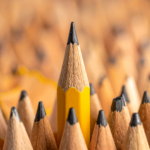 One sharpened yellow pencil rises above a field of near-identical pencils, standing out through its height and precision. The image accompanies an article about perfectionism in the workplace.