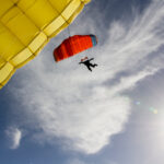 A skydiver in silhouette descends beneath an open red-orange parachute against a bright sunlit sky, framed by the edge of an open yellow parachute in the foreground. The image accompanies an article about risk-taking behavior in leadership.